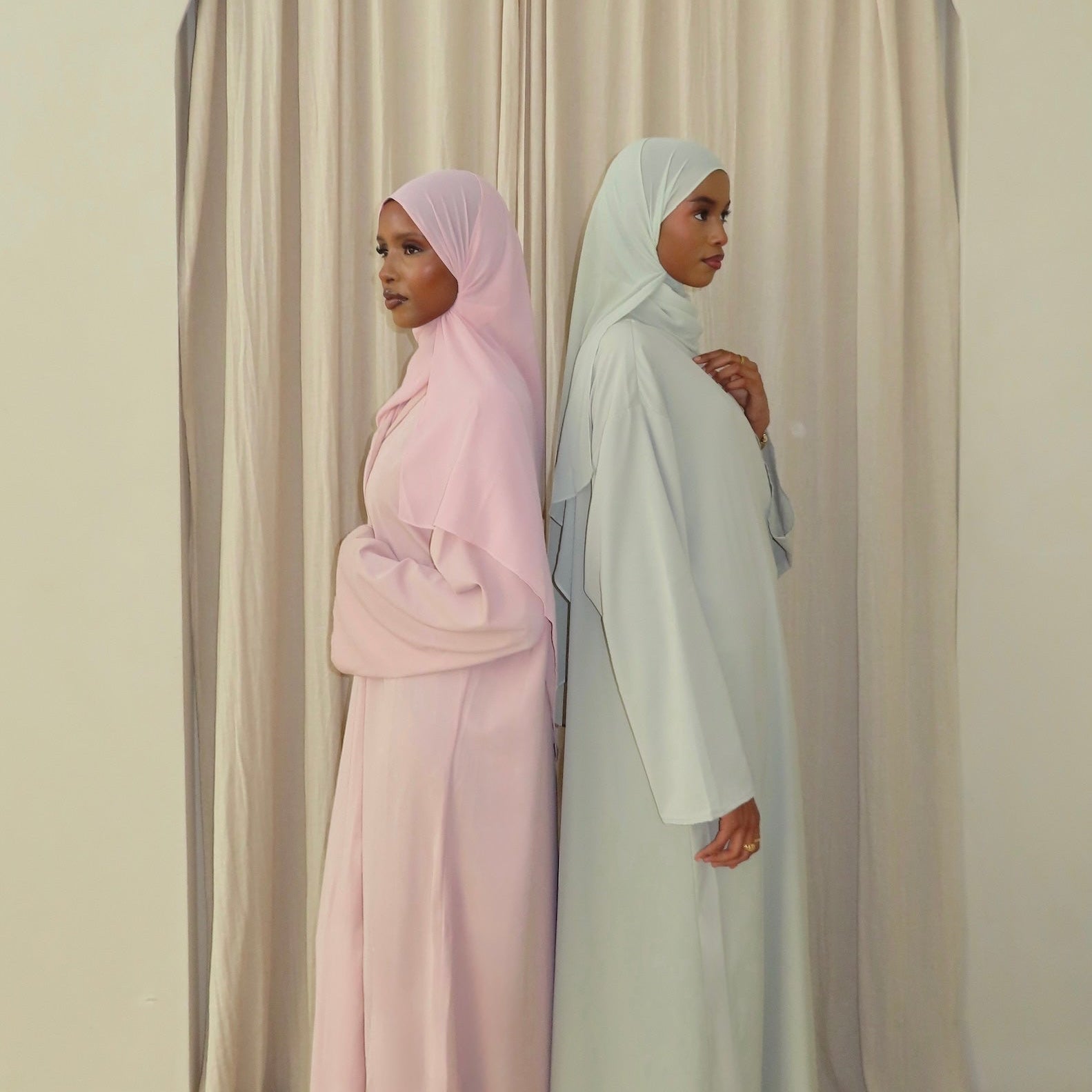Two women wearing modest long abayas and hijabs stand back-to-back in front of draped beige curtains. One abaya is light blush pink, and the other is a very pale icy blue.